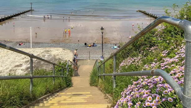 Une plage avec un escalier descendant vers le rivage, une jetée s’avançant dans l’eau et une rambarde au premier plan.