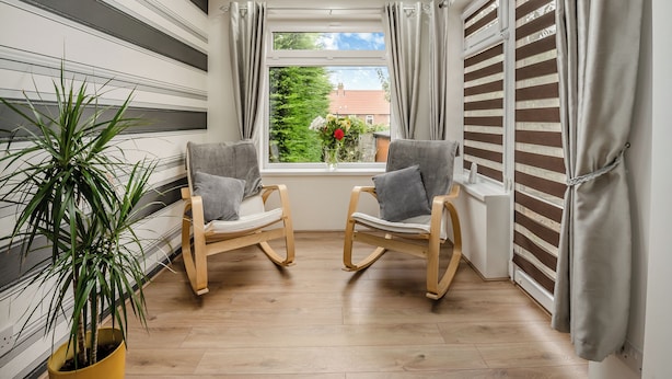 A room with two rocking chairs, a potted plant, a window with curtains, and a view of greenery outside.