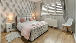 A bedroom with a tufted bed, bedside tables with lamps, a radiator, a window with curtains, and a chair.