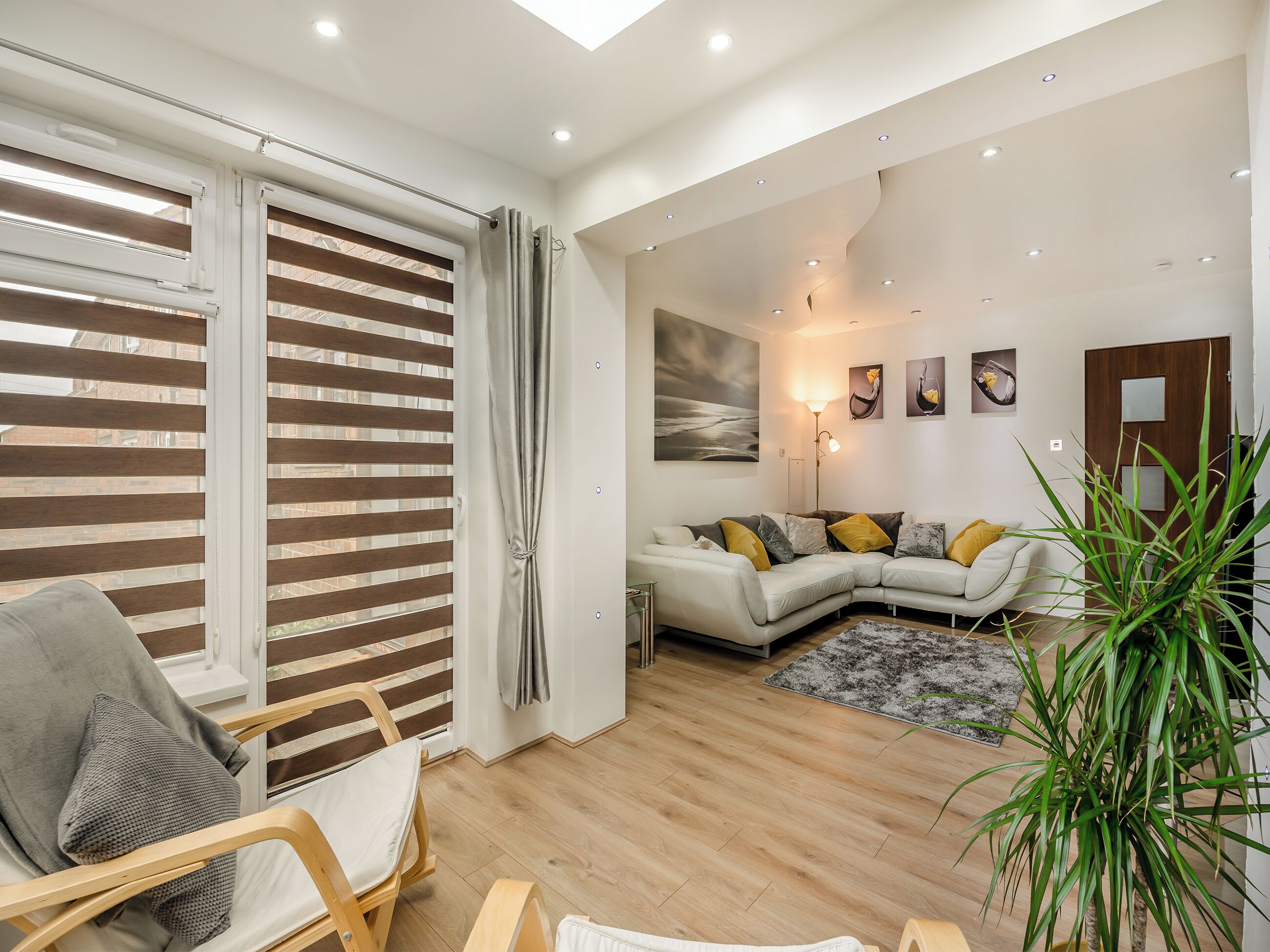 A modern living room with a sectional sofa, a wooden floor, a large window with blinds, and a potted plant.
