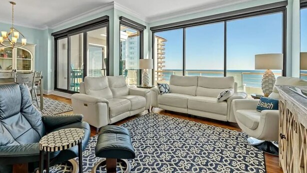 A spacious living room with a large window offering a view of the ocean, a patterned rug, and a mix of modern and classic furniture. - Ocean Creek Penthouse North Myrtle Beach (Myrtle Beach)
