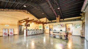 A spacious indoor area with a polished concrete floor, wooden walls, and a high ceiling with metal beams. There are several seating areas with chairs and tables, a refrigerator, and a pool table. - Pet-Friendly Eagletown Home ~ 3 Mi to Kayaking! (Eagletown)