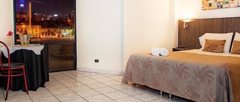 Habitación de hotel con cama, una pequeña mesa con silla y vista a un paisaje urbano nocturno.