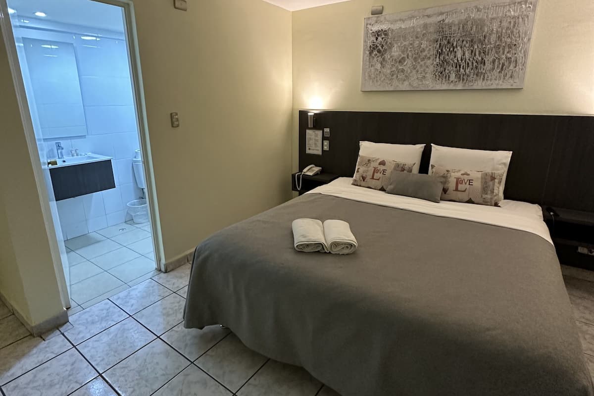 Une chambre d’hôtel avec un lit, une table de chevet, une œuvre d’art accrochée au mur et une salle de bain visible à travers une porte ouverte.