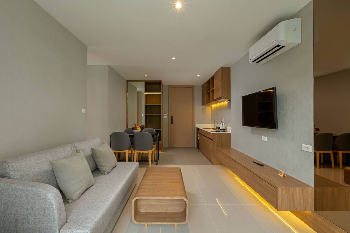 Una sala de estar moderna con un sofá gris, una mesa de centro de madera y una cocina con armarios de madera y un televisor empotrado.