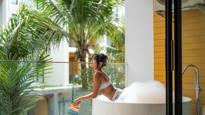 Une femme dans une baignoire dotée d’une porte en verre, entourée de plantes d’intérieur et d’un mur jaune.