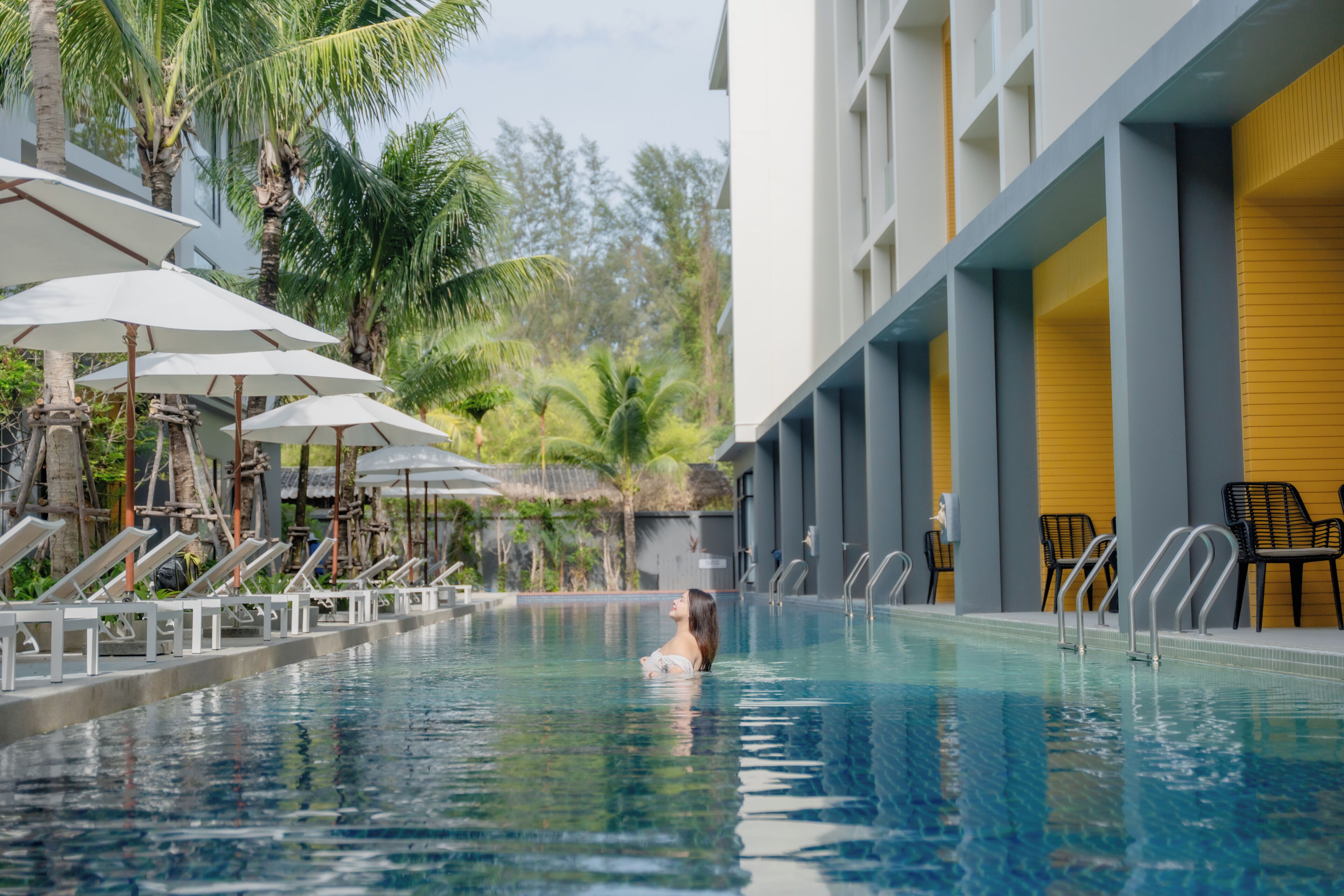 Una piscina con sillas de descanso y sombrillas, rodeada de palmeras y un edificio de fachada amarilla.
