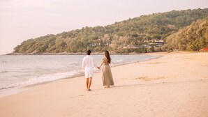 Ein Paar spaziert am Strand mit Blick auf eine Hügelkette und Gebäude im Hintergrund.