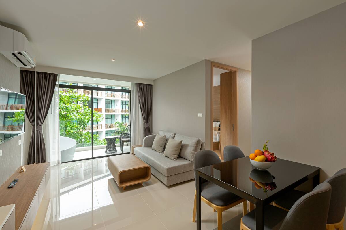 Una sala de estar moderna con un sofá, una mesa de comedor con sillas, un ventanal con cortinas y vistas a un edificio con balcones.