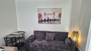 A living room with a grey sofa, a small table, and a chair. A painting of a bridge is on the wall. - Cosy pub flat in Shotts between Glasgow and Edinburgh (Shotts)