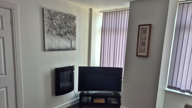 A living room with a flat-screen TV on a black stand, a painting on the wall, and a window with blinds. - Cosy pub flat in Shotts between Glasgow and Edinburgh (Shotts)