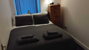 A neatly made bed with a dark bedspread and two pillows, a wooden dresser with a lamp, and a window with blue curtains. - Cosy pub flat in Shotts between Glasgow and Edinburgh (Shotts)