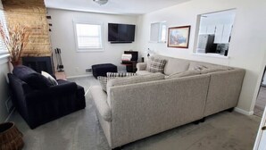 Una sala de estar con un sofá beige grande, un sillón oscuro y un banquito negro pequeño. Un televisor de pantalla plana está montado en la pared, y hay una chimenea con un jarrón decorativo a la izquierda.