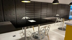 A modern waiting area with a black cushioned bench, a glass-top table, and white chairs with metal legs. - Elegance Suítes Carvalho (São Paulo)