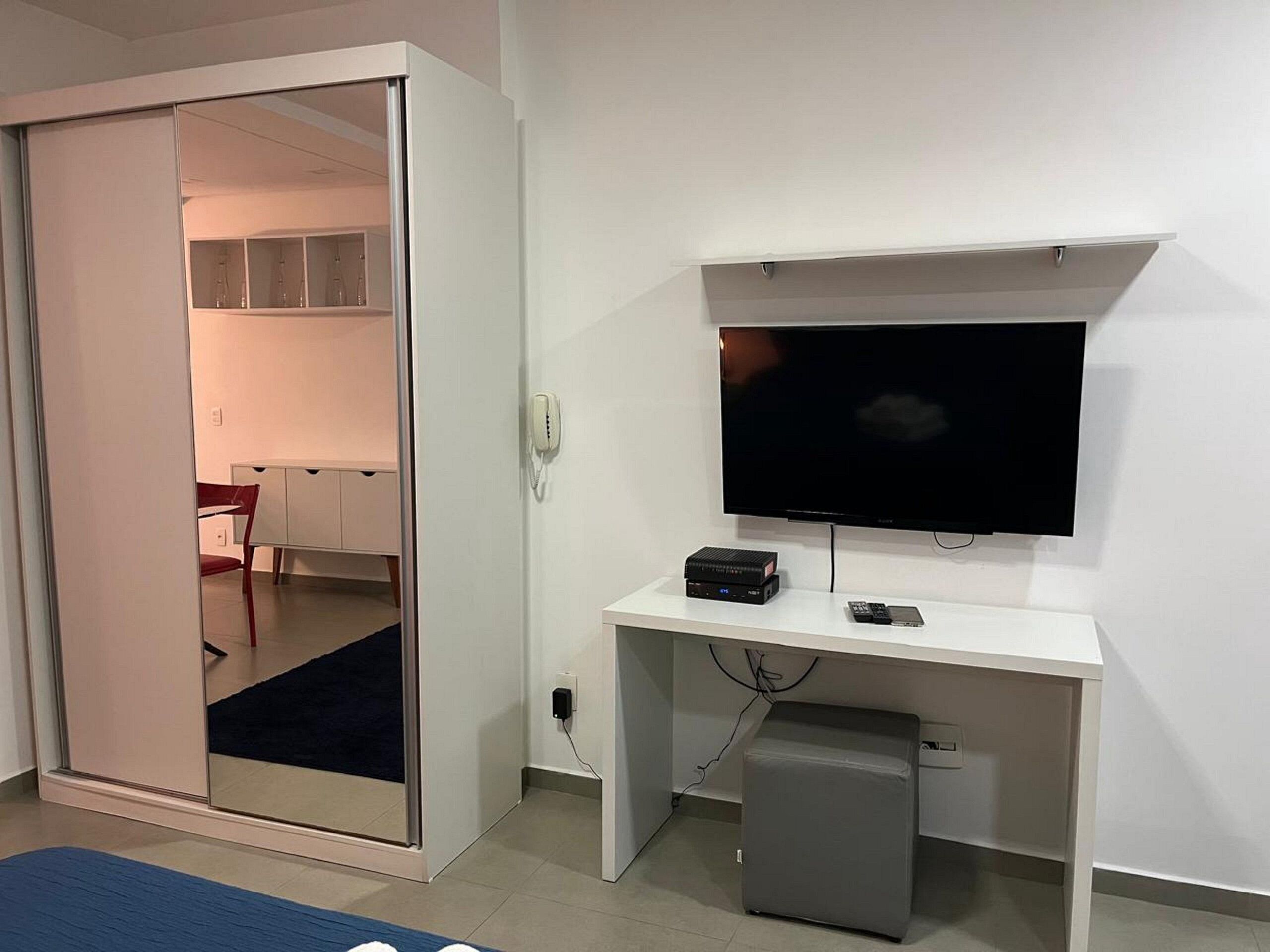 Una habitación con una puerta corredera que da a una cocina, un televisor de pantalla plana montado sobre un escritorio blanco y un refrigerador pequeño debajo del escritorio.