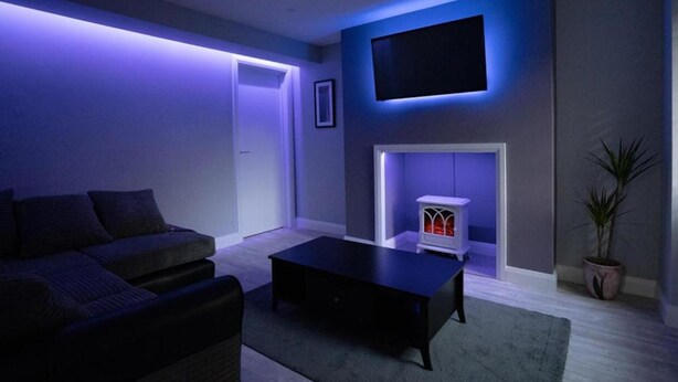 A modern living room with a sofa, a coffee table, a flat-screen TV mounted on the wall, and a fireplace with a white mantel. - Indigo Apartment - Beautiful 1-bed in Ballycastle (Ballycastle)