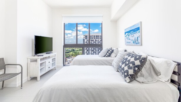 Quarto com cama, televisão em um armário branco, uma cadeira e uma janela grande com vista para a cidade.
