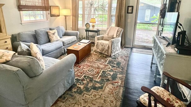 Sala de estar con sofá gris, alfombra estampada, ventilador de techo y vistas al exterior a través de un ventanal.