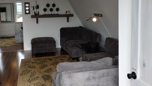 En moderne stue med loft til kip, grå sofaer, trægulv og et spiseområde, der ses igennem en åben dør.
