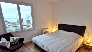 Ein Schlafzimmer mit einem Bett, einem Sessel, einem Nachttisch und einem Fenster mit Blick auf Gebäude und das Meer.