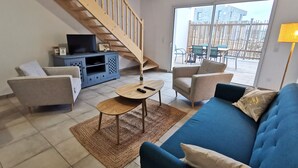 Ein Wohnzimmer mit einem blauen Sofa, einer Holztreppe, einem Fernsehschrank und Blick ins Freie.