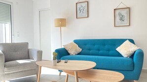 Ein Wohnzimmer mit einem blauen Sofa, einem grauen Sessel und einem runden Holztisch. An der Wand hängen zwei gerahmte Kunstwerke und es steht eine Stehlampe im Raum.