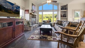 A spacious living room with a stone fireplace, a large flat-screen TV, and a view of a green landscape through large windows. - Cozy Cottage Inside the Premier Old Kinderhook Golf Resort (Camdenton)