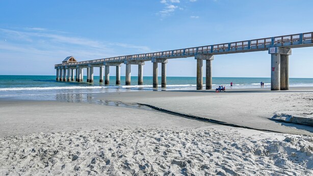 A pier extending into the ocean with a building at the end, a sandy beach, and people in the distance.