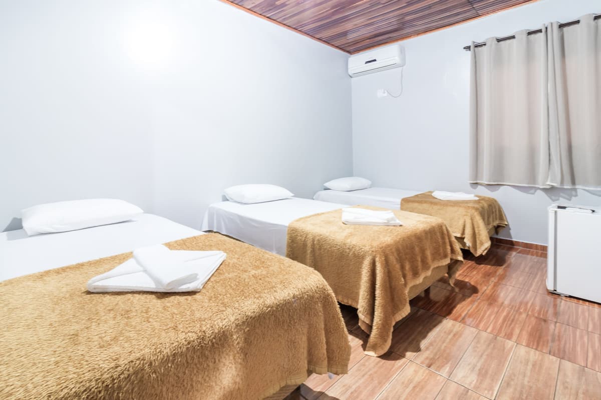 Ein Hotelzimmer mit drei Betten, jedes mit einem weißen Kopfkissen und einer braunen Decke. Auf der rechten Seite des Zimmers steht ein kleines, weißes Gerät.