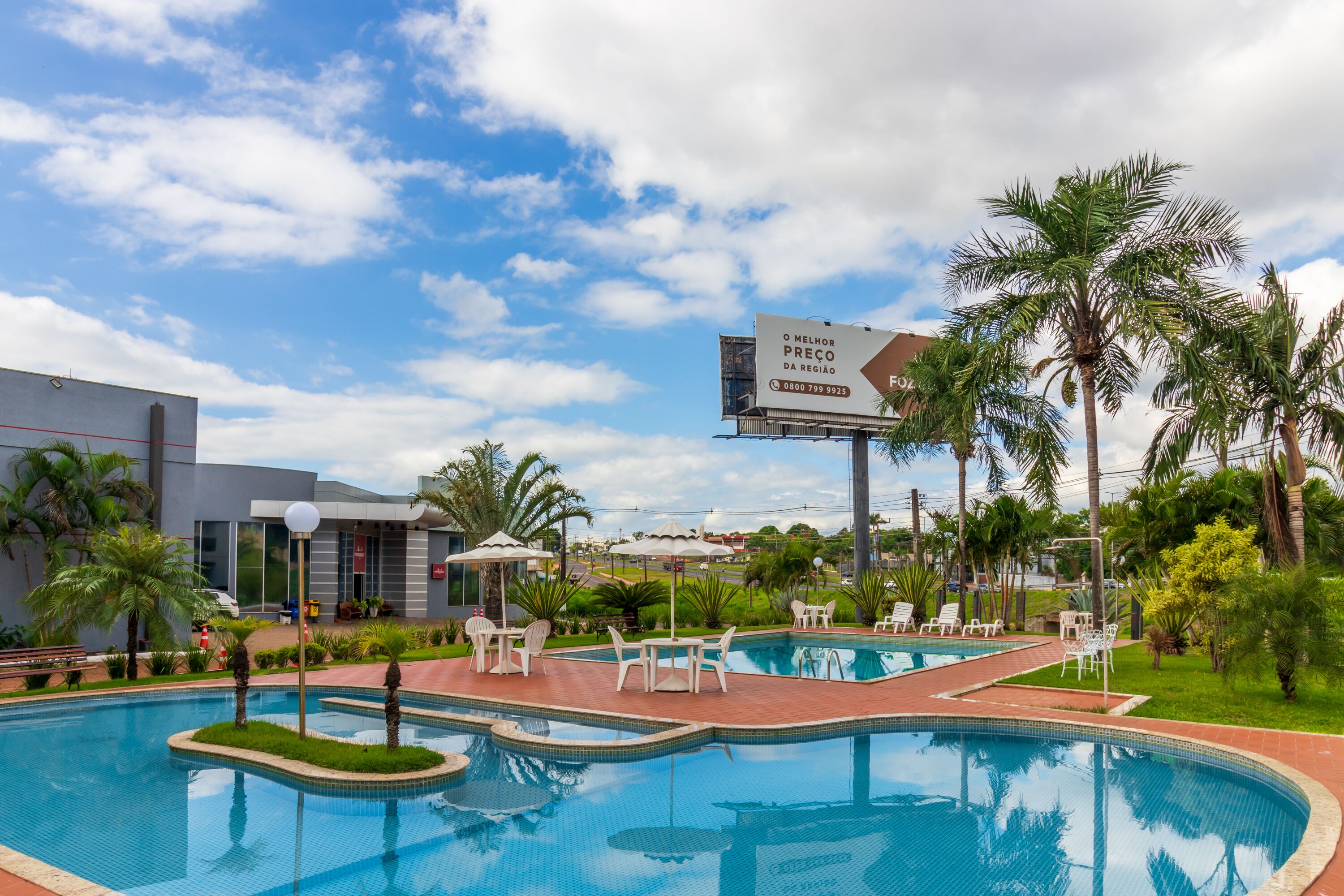 Una piscina con sillones de descanso y sombrillas, rodeada de palmeras y un edificio con un cartel.
