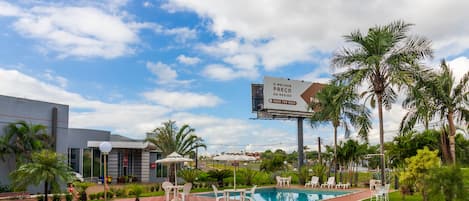 Una piscina con sillones de descanso y sombrillas, rodeada de palmeras y un edificio con un cartel.