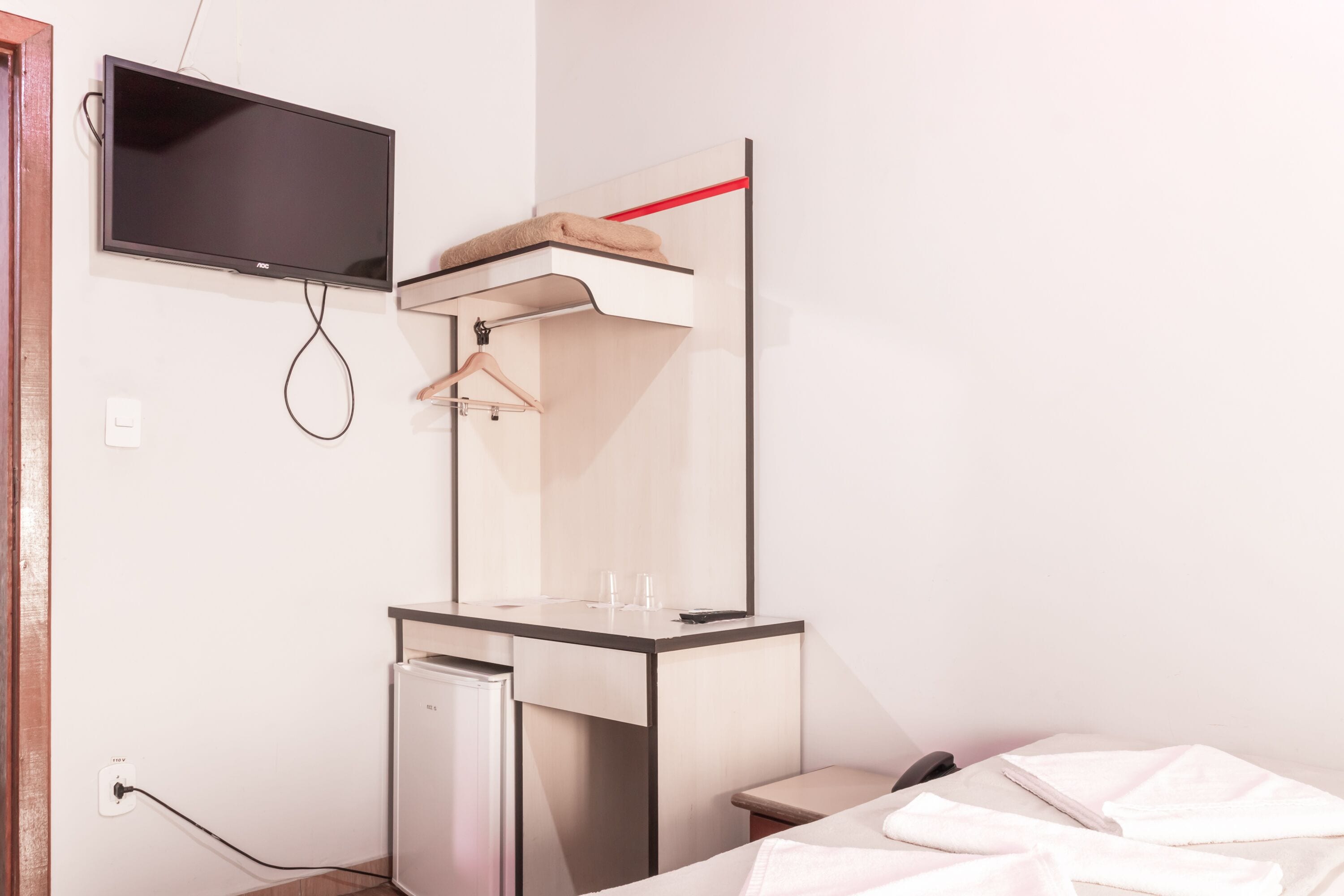 A hotel room with a bed, a desk with a mirror, a television mounted on the wall, and a small refrigerator.