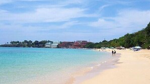 A beach with clear turquoise water, a sandy shore, and buildings in the background.