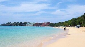 A beach with clear turquoise water, a sandy shore, and buildings in the background. - Luxury Beachfront Condo - Hispaniola Beach 5B3 OceanView / 2 bed 2.5 bath (Sosúa)