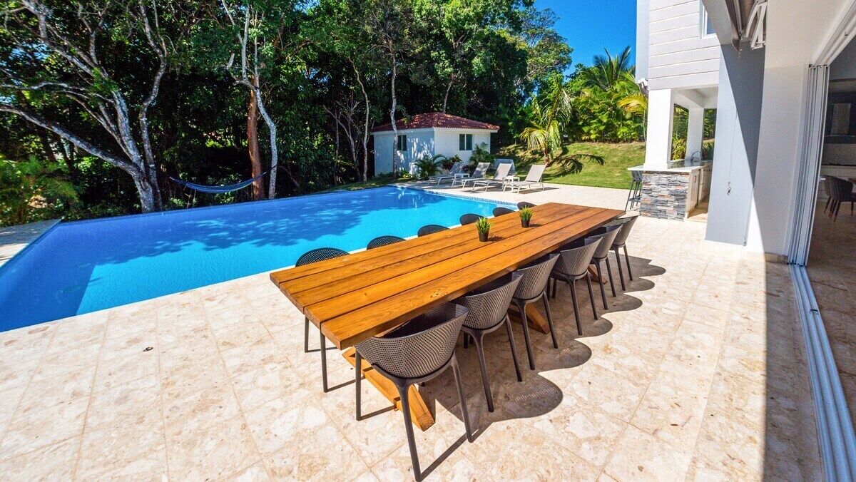 Ein Poolbereich mit Holztisch und -stühlen, einer Terrasse mit Steinplatten und Blick auf ein Haus und Bäume.