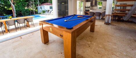 A pool table in a spacious room with a view of a swimming pool and outdoor seating.