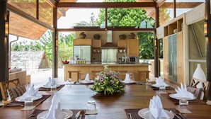 Un coin repas avec une table en bois dressée pour le repas, entourée de chaises en bois, et offrant une vue sur la cuisine et la verdure environnante.