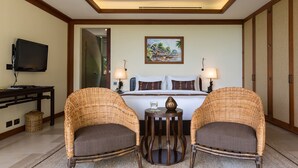 Une chambre d’hôtel avec un lit, deux fauteuils en osier, une petite table, un téléviseur et un tableau accroché au mur.