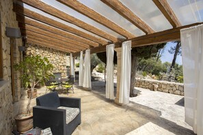 Een terras buiten met stenen muren, een pergola en een zithoek met een tafel en stoelen.