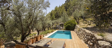 A wooden deck with a pool, surrounded by olive trees and a stone wall.