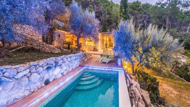 A swimming pool with a stone wall and a wooden deck, surrounded by trees and a stone building in the background. - 3 bedroom villa with private pool (Mancor de la Vall)