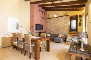 A cozy living room with a stone wall, a fireplace, a television, and a dining area with wicker chairs and a wooden table. - 3 bedroom villa with private pool (Mancor de la Vall)