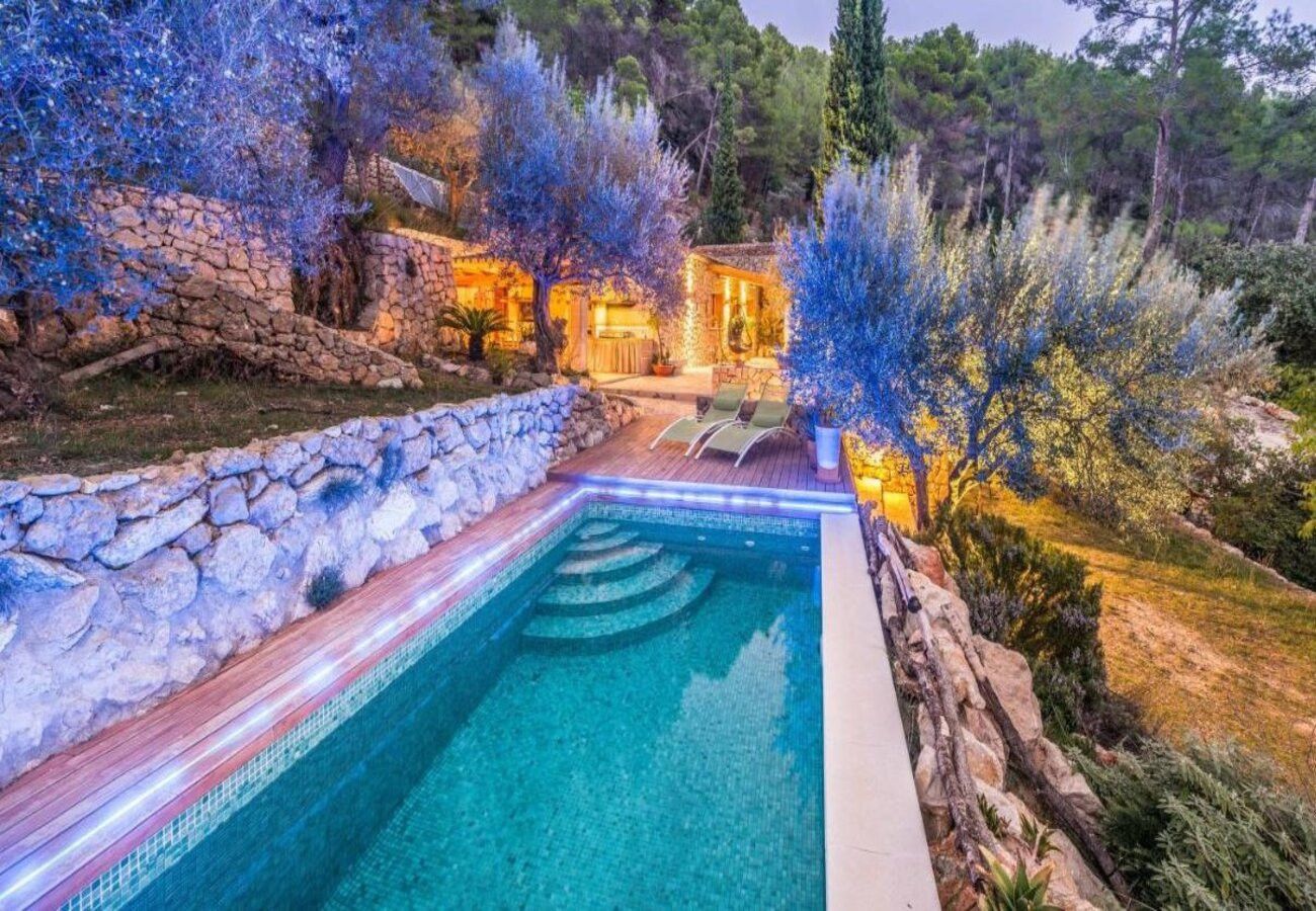 Une piscine bordée d’un mur en pierre et dotée d’une terrasse en bois, entourée d’arbres et avec un bâtiment visible au loin.