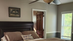 Un dormitorio con una cama de madera oscura, un ventilador de techo, un cuadro en la pared y una ventana con persianas.