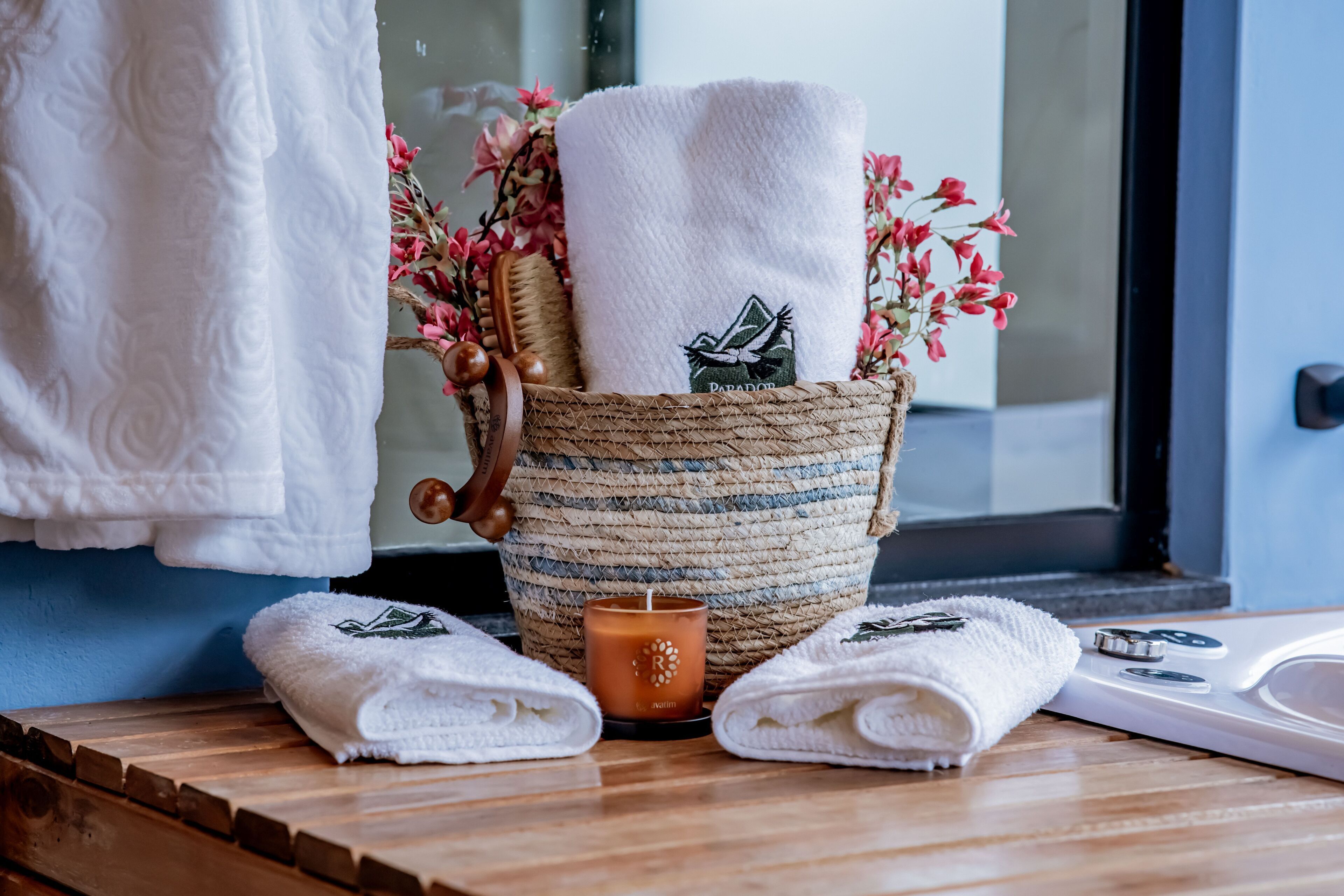 Un baño con tocador de madera, una toalla blanca, una cesta de mimbre con toallas y una vela.