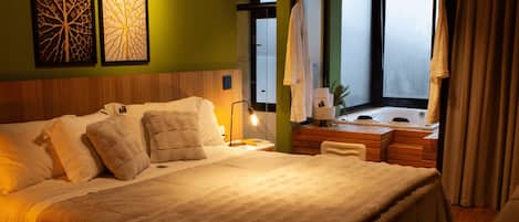 Habitación de hotel con una cama grande, cabecera de madera, mesita de noche con lámpara, tocador con espejo y ventana con cortinas.