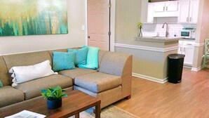 A living room with a sectional sofa, a coffee table with a plant, and a kitchen area with white cabinets and a microwave.