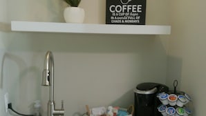 A kitchen with a granite countertop, a stainless steel sink, a coffee maker, a basket with kitchen utensils, and a sign above the counter.