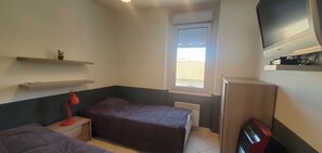 A small, compact room with two beds, a desk, a red desk lamp, a TV mounted on the wall, and a window with blinds. - Sea view and 50m from the beaches, 3-room apartment on the 1st floor of a villa, air-conditioned and a beautiful terrace for 4 privileged people (Fréjus)