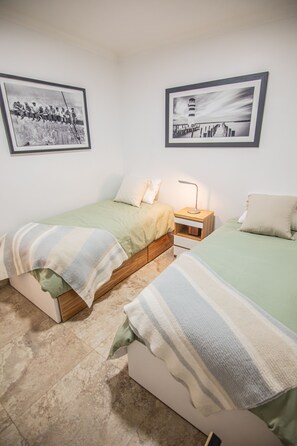 Un dormitorio con dos camas individuales, una mesita de noche con lámpara y dos cuadros enmarcados en la pared.